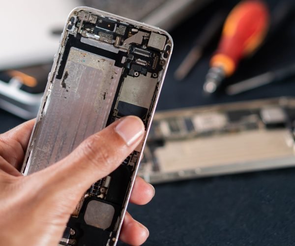A close-up of a person holding a disassembled smart phone, revealing its internal components. The background features tools and other electronic devices, indicating a repair or assembly workspace.