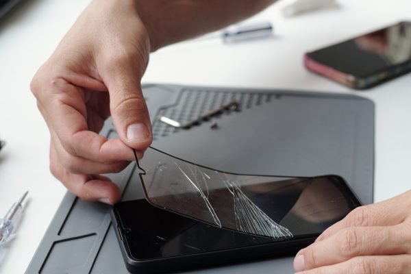 Close-up of hands repairing cracked smartphone screen using specialized tools on tech workbench. Many small components and electronic devices are scattered around