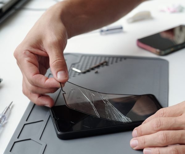 Close-up of hands repairing cracked smartphone screen using specialized tools on tech workbench. Many small components and electronic devices are scattered around